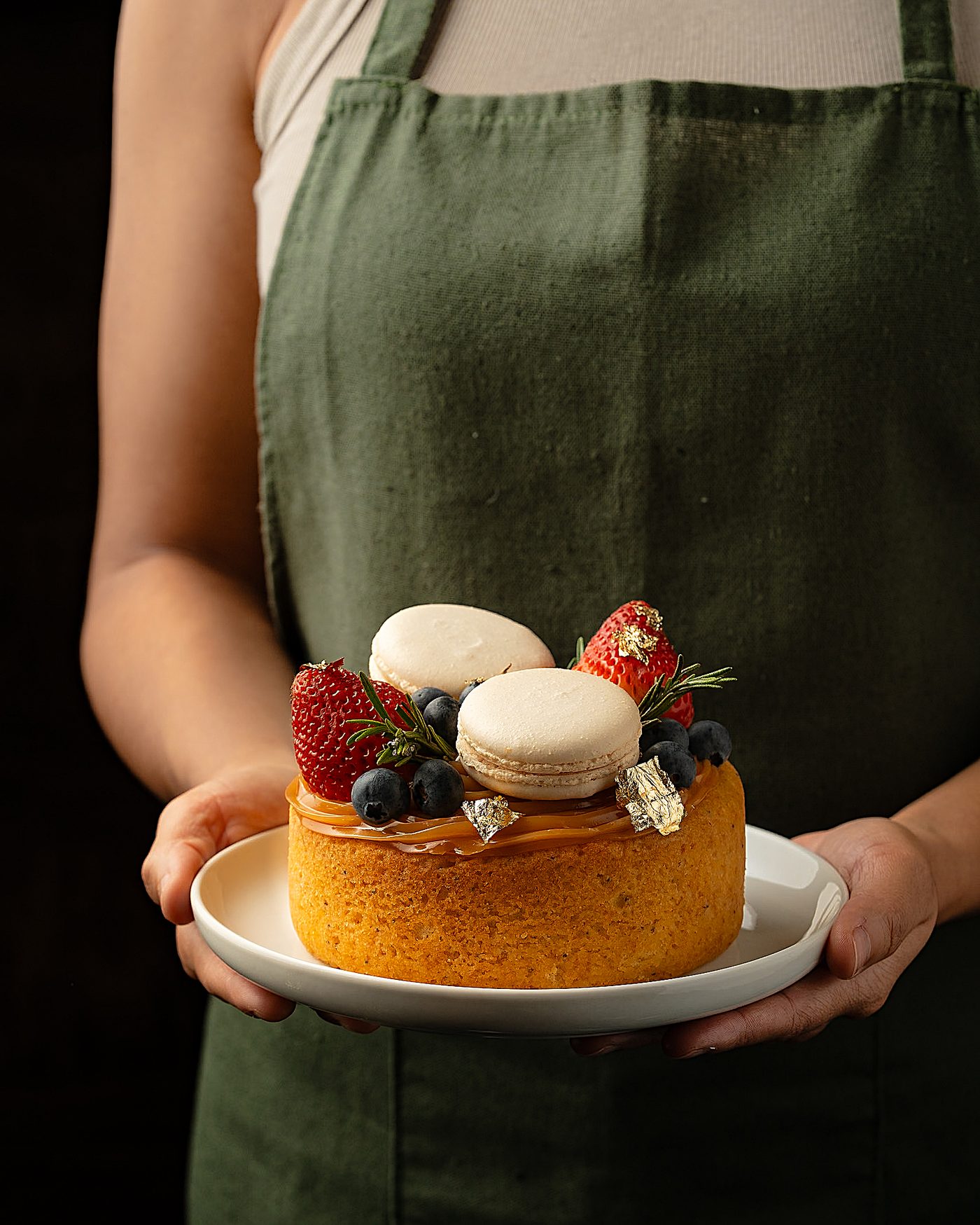 Torta con macarons, frutos rojos y hoja de oro sostenida por un pastelero con delantal verde